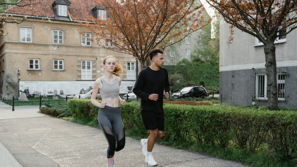 Two adults jogging on a sidewalk in an urban setting surrounded by spring foliage.